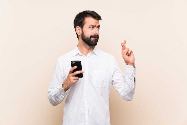 Junger mann mit dem bart, der ein mobile mit den fingern kreuzen und das beste wünschen hält