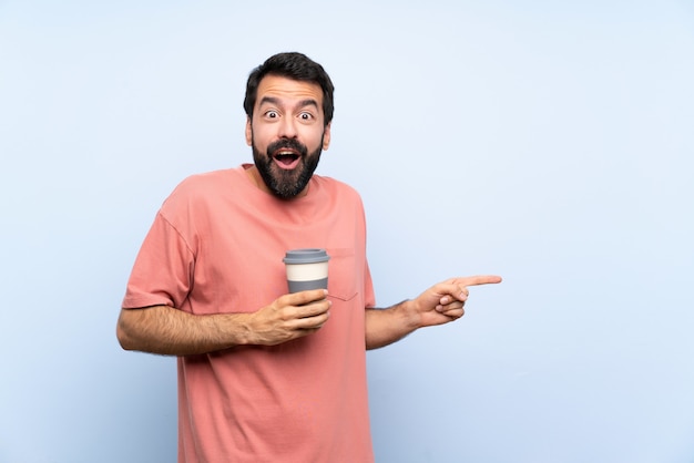 Junger Mann mit dem Bart, der einen Mitnehmerkaffee auf Blau hält, überraschte und Finger auf die Seite zeigte