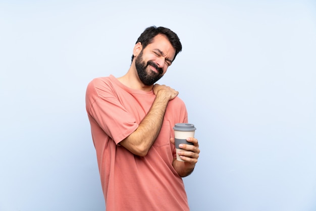 Junger Mann mit dem Bart, der einen Mitnehmerkaffee über dem lokalisierten Blau leidet unter den Schmerz in der Schulter hält