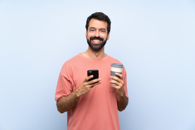 Junger Mann mit dem Bart, der einen Mitnehmerkaffee über der lokalisierten blauen Wand sendet eine Mitteilung mit dem Mobile hält