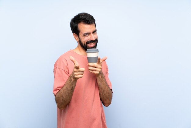 Junger Mann mit dem Bart, der einen Mitnehmerkaffee über der lokalisierten blauen Wand zeigt auf die Front und das Lächeln hält