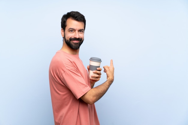 Junger Mann mit dem Bart, der einen Mitnehmerkaffee über der lokalisierten blauen Wand zurück zeigt hält