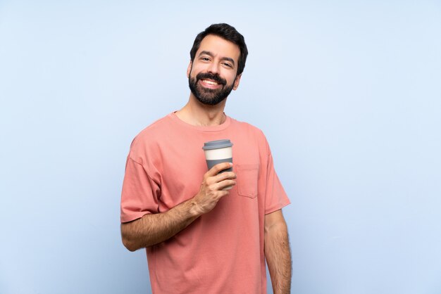 Junger Mann mit dem Bart, der einen Mitnehmerkaffee über getrenntem blauem Lachen anhält