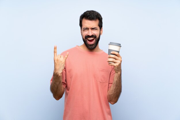 Junger Mann mit dem Bart, der einen Mitnehmerkaffee über lokalisiertem Blau macht Felsengeste hält