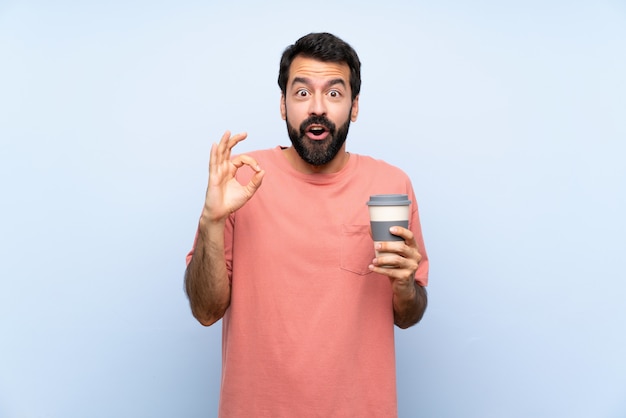 Junger Mann mit dem Bart, der einen Mitnehmerkaffee über lokalisiertem Blau überrascht und okayzeichen zeigend hält
