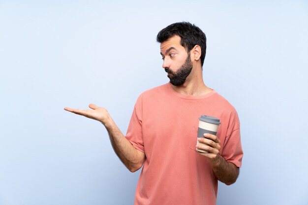 Junger Mann mit dem Bart, der einen Mitnehmerkaffee über lokalisiertem blauem haltenem copyspace mit Zweifeln hält