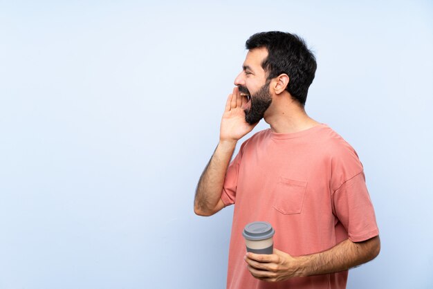 Junger Mann mit dem Bart, der einen Mitnehmerkaffee über lokalisiertem blauem Schreien mit dem breiten Mund hält, öffnen sich