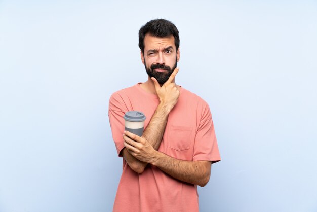 Junger Mann mit dem Bart, der einen Mitnehmerkaffee über lokalisiertem blauem Wanddenken hält