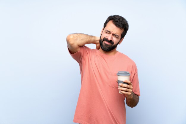 Junger Mann mit dem Bart, der einen Mitnehmerkaffee über lokalisierter blauer Wand mit Halsschmerzen hält