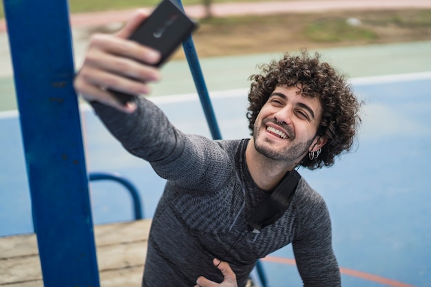 Foto junger mann mit dem lockigen haar, das nach sport sitzt und ein selfie mit einer covid-19-maske nimmt