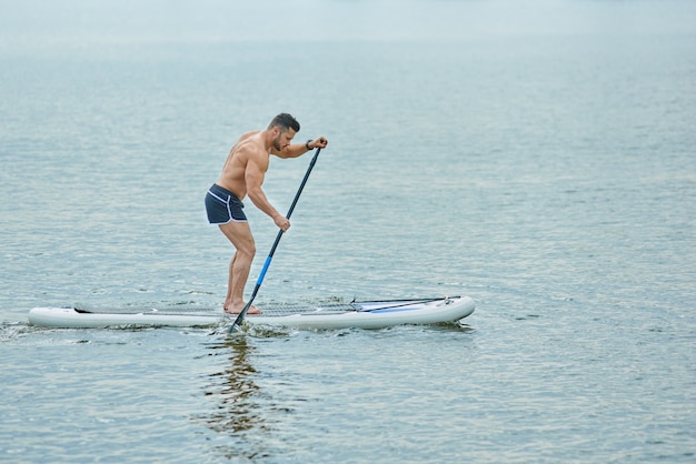 Junger Mann mit dem sportlichen Körper, der auf dem Supbrett, durchstreifend mit einer Pfütze steht.