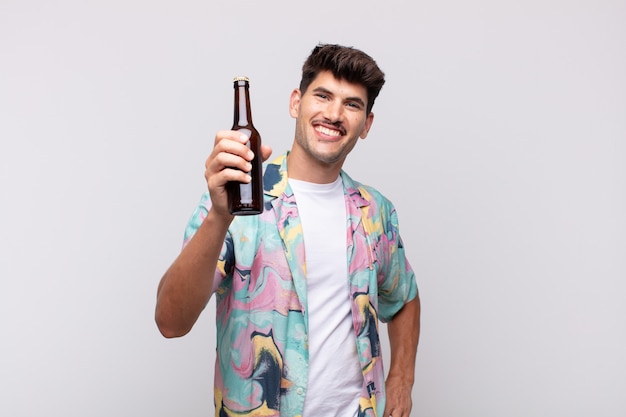 Junger Mann mit einem Bier, das glücklich mit einer Hand auf Hüfte und selbstbewusster, positiver, stolzer und freundlicher Haltung lächelt