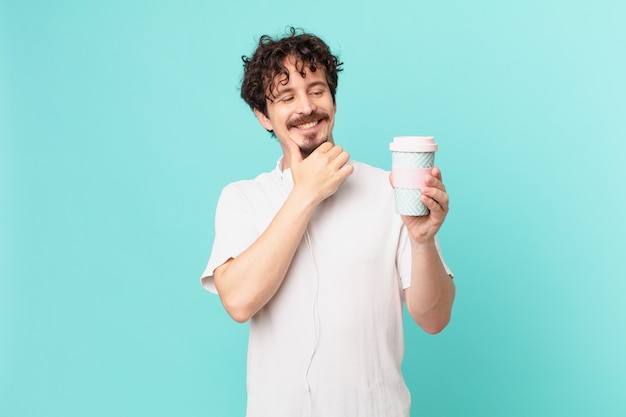 Junger Mann mit einem Kaffee, der mit einem glücklichen, selbstbewussten Ausdruck mit der Hand am Kinn lächelt