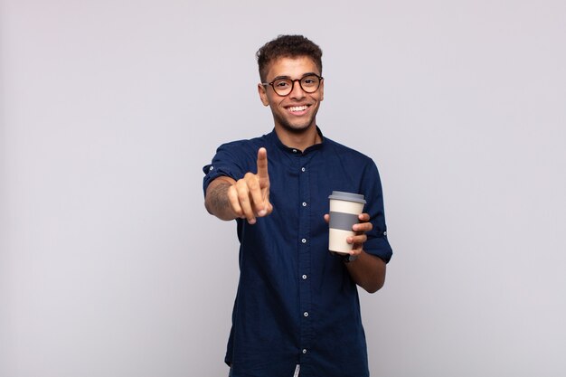 Junger Mann mit einem Kaffee, der stolz und zuversichtlich lächelt und Nummer eins triumphierend posiert und sich wie ein Anführer fühlt