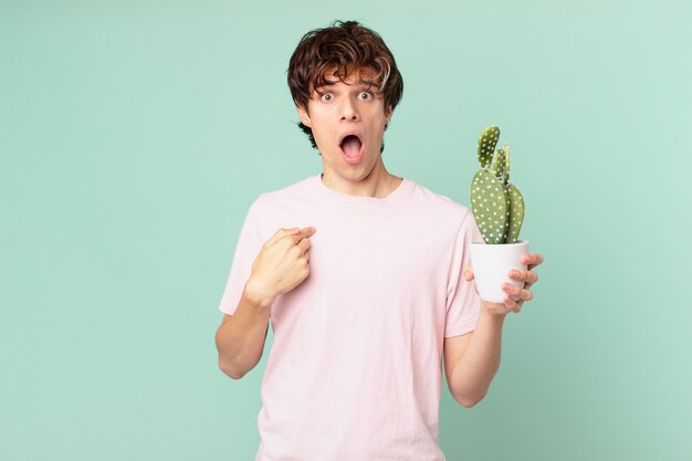 Junger Mann mit einem Kaktus, der schockiert und überrascht mit weit geöffnetem Mund aussieht und auf sich selbst zeigt