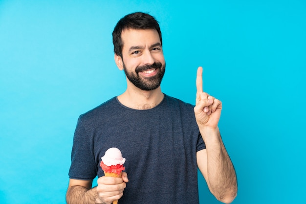 Junger Mann mit einem Kornetteis über der isolierten blauen Wand, die einen Finger im Zeichen des Besten zeigt und hebt