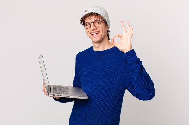 Junger Mann mit einem Laptop, der sich glücklich fühlt und Zustimmung mit einer okayen Geste zeigt
