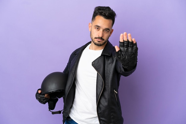 Junger Mann mit einem Motorradhelm auf violettem Hintergrund isoliert, der Stoppgeste macht