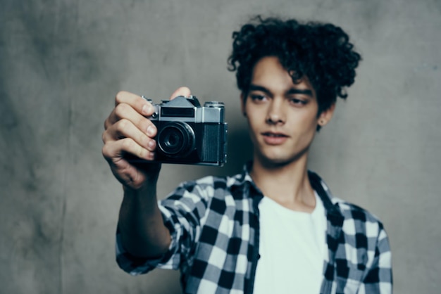 Junger Mann mit einer Kamera in der Hand und in einem karierten T-Shirt auf grauem Hintergrund drinnen Fotograf