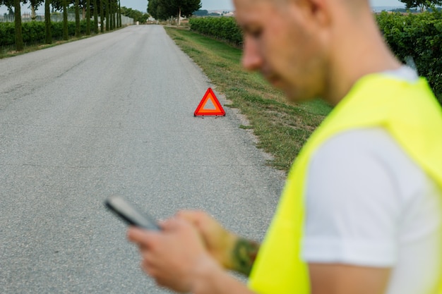 Junger Mann mit gelber reflektierender Weste seine Autounterstützung nah an seinem defekten Auto anrufend