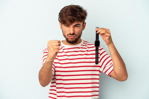 Junger Mann mit gemischter Rasse, der eine Armbanduhr auf blauem Hintergrund hält, die Faust zur Kamera zeigt, aggressiver Gesichtsausdruck.