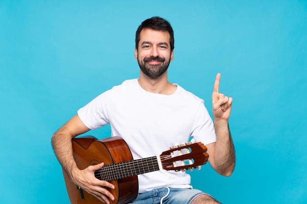 Junger Mann mit Gitarre über dem lokalisierten Blau, das einen Finger im Zeichen des Besten zeigt und anhebt