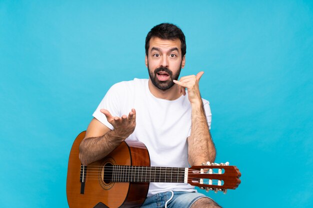 Junger Mann mit Gitarre über der lokalisierten blauen Wand, die Telefongeste und -c $ zweifeln macht