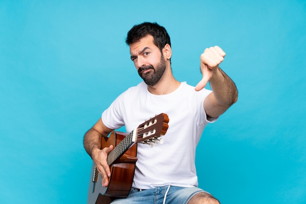 Junger Mann mit Gitarre über der lokalisierten blauen Wand, die unten Daumen mit negativem Ausdruck zeigt