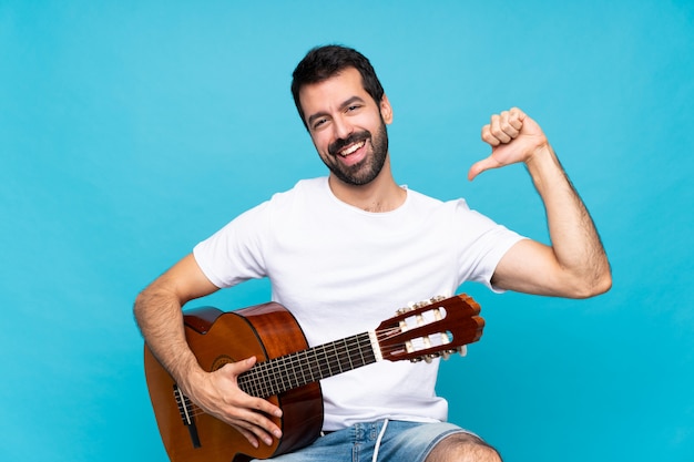 Junger Mann mit Gitarre über getrenntem Blau stolz und selbstzufrieden