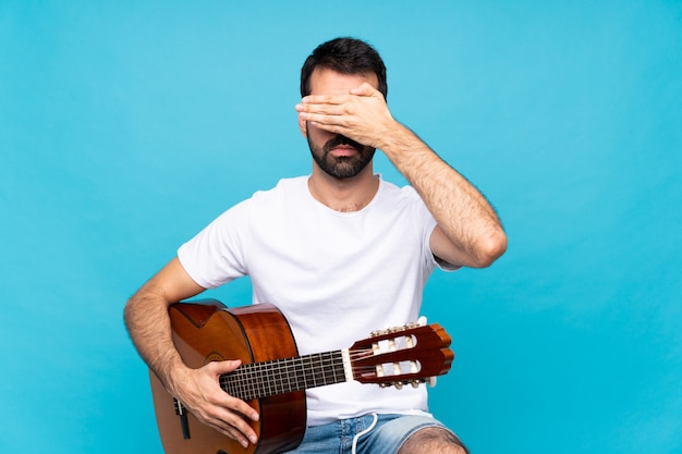 Junger Mann mit Gitarre über lokalisierten blauen Bedeckungsaugen durch Hände, möchten nicht etwas sehen