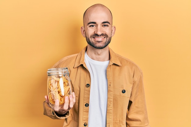 Junger Mann mit Glatze, der ein Glas mit ungekochten Nudeln in der Hand hält, sieht positiv und glücklich aus und lächelt mit einem selbstbewussten Lächeln, das Zähne zeigt