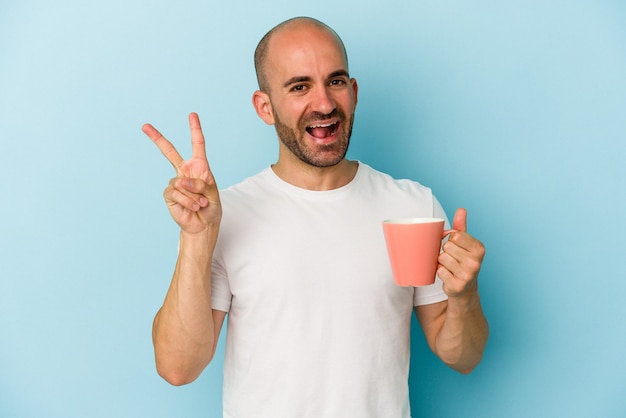 Junger Mann mit Glatze, der einen Becher auf blauem Hintergrund isoliert hält, freudig und sorglos, der ein Friedenssymbol mit den Fingern zeigt.