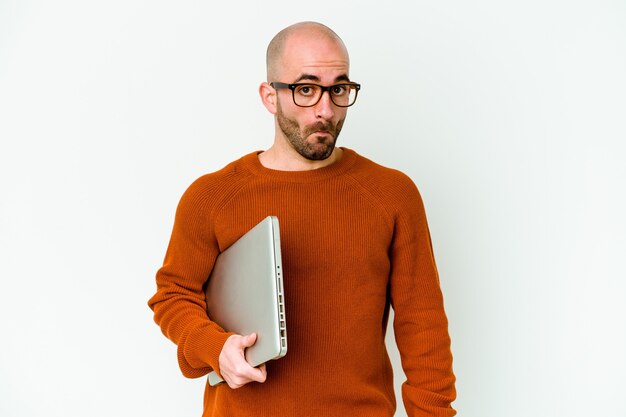 Junger Mann mit Glatze, der einen Laptop isoliert auf weißem Hintergrund hält, zuckt mit den Schultern und öffnet die Augen verwirrt.