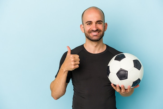 Junger Mann mit Glatze, der Fußball spielt, isoliert auf blauem Hintergrund, lächelnd und Daumen hoch