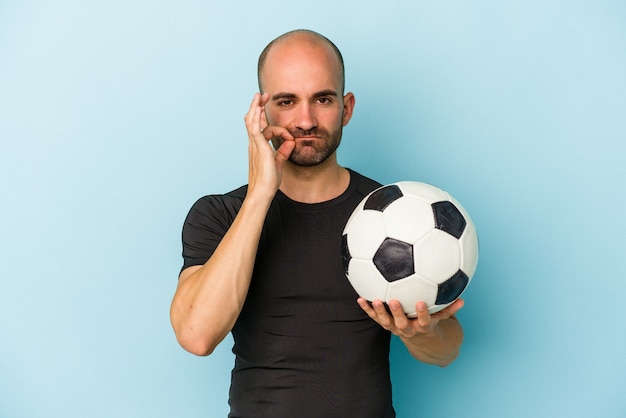 Junger Mann mit Glatze, der Fußball spielt, isoliert auf blauem Hintergrund mit den Fingern auf den Lippen, die ein Geheimnis halten.