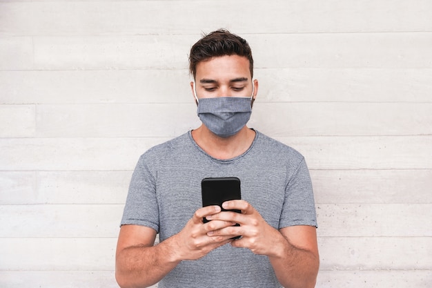 Junger Mann mit Handy-App beim Tragen einer Gesichtsschutzmaske während des Ausbruchs des Coronavirus - Millennial-Typ, der Videos auf dem Smartphone zeigt - Covid 19 Lifestyle- und Technologiekonzept