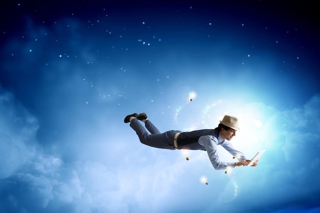Junger Mann mit Hut und Freizeitkleidung, der mit Tablet am Himmel schwebt. Gemischte Medien