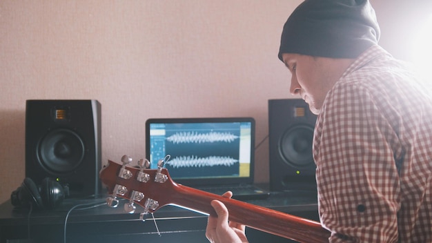 Foto junger mann mit hutmusiker komponiert und nimmt soundtrack auf, der gitarre spielt, mit computer, kopfhörern und tastatur, fokus auf ausrüstung