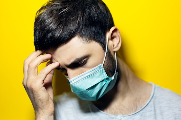 junger Mann mit Kopfschmerzen, Hand auf Kopf haltend, medizinische Grippemaske auf gelber Wand tragend.