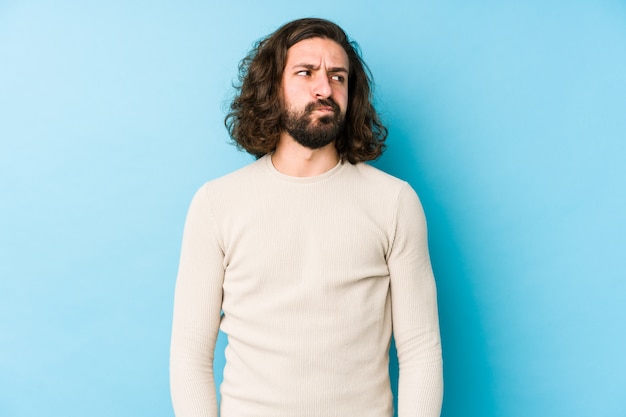 Junger Mann mit langen Haaren an einer blauen Wand verwirrt, fühlt sich zweifelhaft und unsicher.