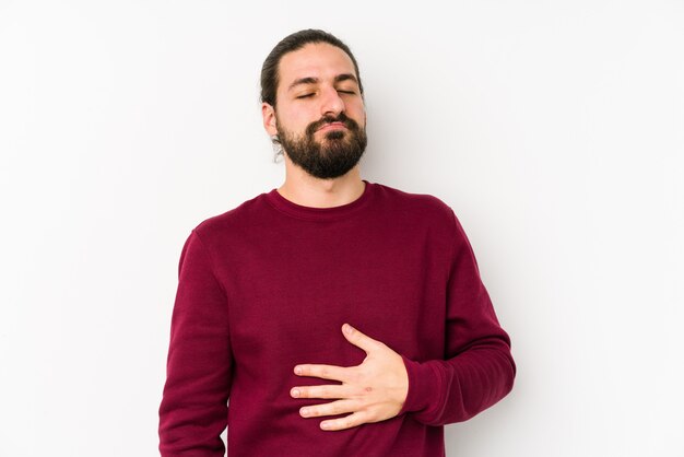 Junger Mann mit langen Haaren an einer weißen Wand berührt den Bauch und lächelt sanft