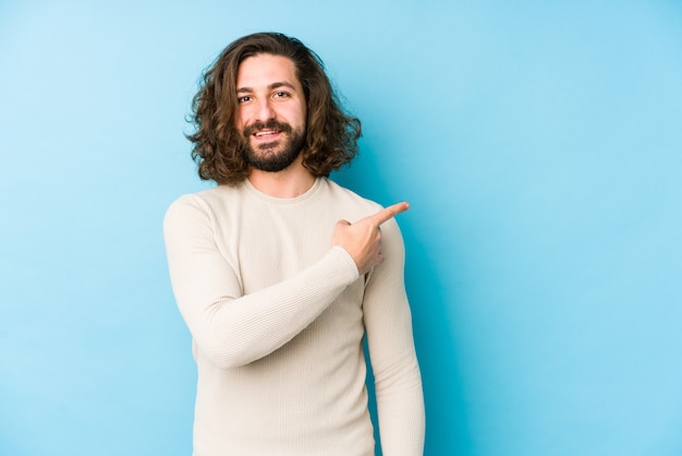 Junger Mann mit langen Haaren auf einer blauen Wand, die lächelt und zur Seite zeigt und etwas an der leeren Stelle zeigt.