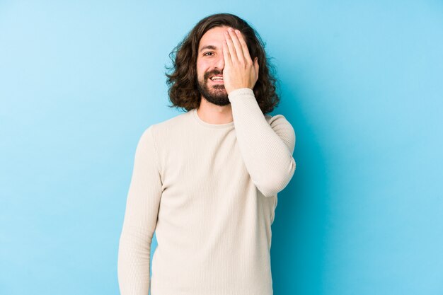 Junger Mann mit langen Haaren auf einer blauen Wand, die Spaß hat, die Hälfte des Gesichts mit Handfläche bedeckend.