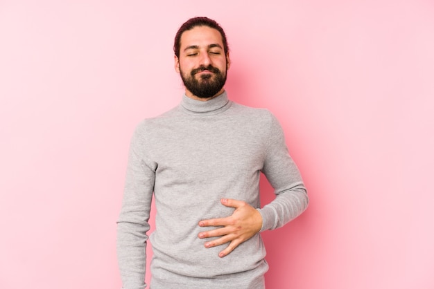 Junger Mann mit langen Haaren, der auf einem rosa Raum isoliert ist, berührt Bauch, lächelt sanft, Ess- und Zufriedenheitskonzept.