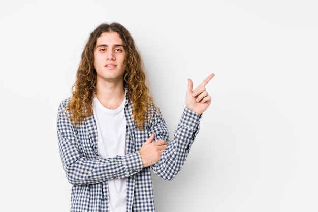 Junger Mann mit langen Haaren, der ein isoliertes Lächeln darstellt, das fröhlich mit Zeigefinger weg zeigt.