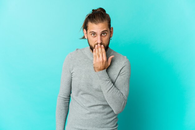 Junger Mann mit langen Haaren sieht schockiert aus, bedeckt den Mund mit den Händen und ist bestrebt, etwas Neues zu entdecken.