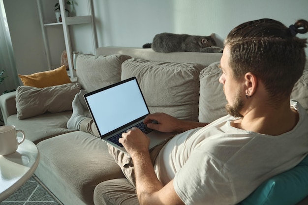 Junger Mann mit leerem Laptop mit leerem Bildschirm, der zu Hause auf der Couch liegt und arbeitet