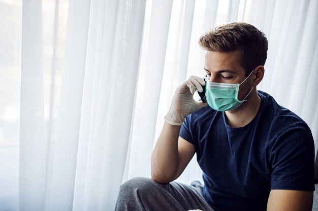 Junger Mann mit Maske und Handschuhen, die Handy tippend betrachten und besorgt schauen. Bleib zu Hause, Weltpandemie. Quarantäne.