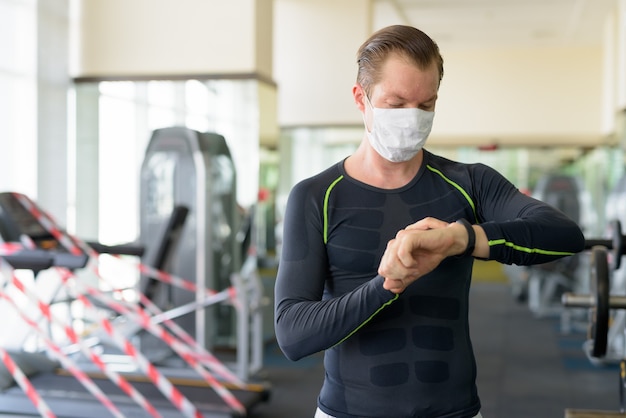 Junger Mann mit Maske zum Schutz vor Coronavirus-Ausbruch, der Smartwatch im Fitnessstudio während Coronavirus covid-19 überprüft