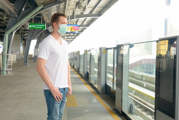 Junger Mann mit Maske zum Schutz vor dem Ausbruch des Koronavirus, der am Bahnhof steht und wartet